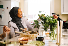 Happy Young Woman Wearing Hijab Passing Food To Friend At Dining Table