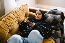 Smiling Young Woman Using Smart Phone Lying On Sofa At Home