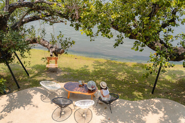 Wall Mural - Beautiful woman wearing hat playing with pet on small wooden coffee table and modern steel chairs in garden near nature river with swing chair under the tree relaxing time on summer holiday vacation.