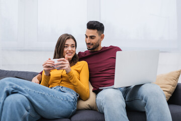 Wall Mural - Cheerful woman holding cup of coffee near arabian boyfriend with laptop on couch.