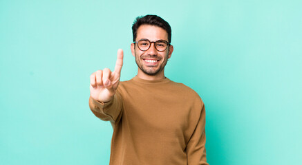 young handsome hicpanic man smiling and looking friendly, showing number one or first with hand forward, counting down