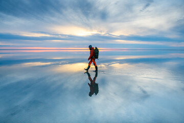 Wall Mural - Man traveler with backpack walking on the salt lake at sunset. Sky with clouds are reflected in the mirror water surface. Travel and adventure concept