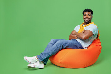 Wall Mural - Full body young man of African American ethnicity wear blue t-shirt sit in bag chair hold hands crossed folded look camera isolated on plain green background studio portrait. People lifestyle concept.