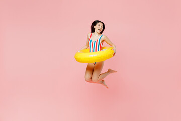 FUll body young woman of Asian ethnicity in striped one-piece swimsuit hawaii lei yellow inflatable ring jump high into water isolated on plain pastel pink background Summer vacation sea rest concept