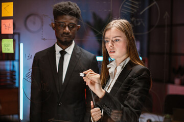 Wall Mural - Team of two professional multicultural traders discussing possible risks and profits on trade market. Male and female partners looking at glass board while standing in modern office conference room.