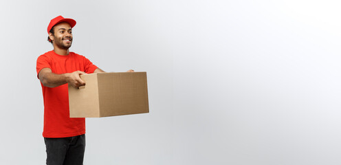 Wall Mural - Delivery Concept - Portrait of Happy African American delivery man in red cloth holding a box package. Isolated on Grey studio Background. Copy Space.