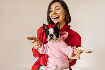 Cheerful young caucasian girl looking at camera, playing with dog holding in her arms indoors. Brunette woman laughs during portrait shoot with french bulldog puppy. Love for pets, joy and tenderness