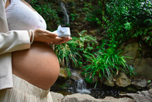 Lady In Her Late Pregnancy Holding Blue Baby Shoes Above Her Belly Outdoors