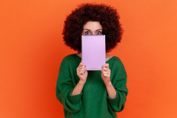 Wall Mural - Portrait of woman with Afro hairstyle wearing green casual style sweater covering half of her face with paper organizer, looking at camera. Indoor studio shot isolated on orange background.