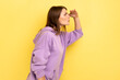 Side view of concentrated attentive beautiful young woman standing seriously, with hand on forehead and looking far, wearing purple hoodie. Indoor studio shot isolated on yellow background.