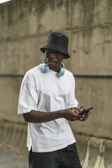 Wall Mural - Young cool black male in a street style outfit with headphones on his neck walking with his phone