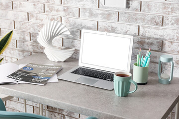 Modern computer, magazines, cup of tea and decor on table near light brick wall