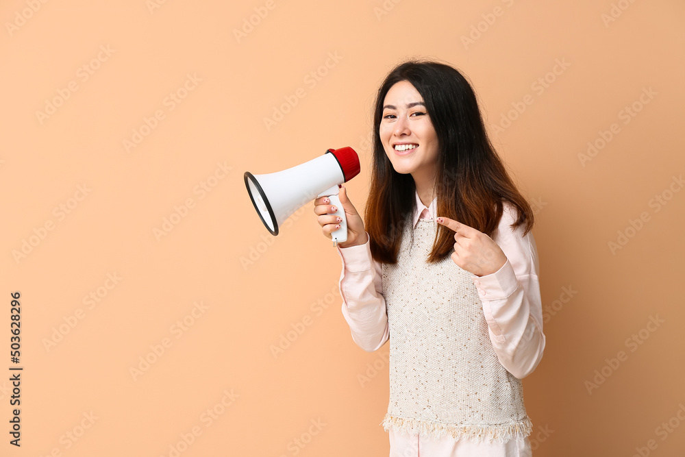 Young Asian woman with megaphone pointing at something on beige background