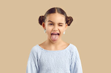 Happy Child Sticking Tongue Out. Studio Shot Of A Cheerful Naughty Kid Making A Funny Grimace. Pretty Little Girl With Adorable Space Hair Buns And Modern Earrings Shows Her Tongue And Winks Her Eye