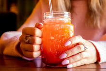Young Caucasian Woman Holds Glass With Handle Of Red Strawberry Lemonade With Ice On Wooden Table. Female Hands With Pink Manicure And Cocktail. Front View, Closeup, Lifestyle.