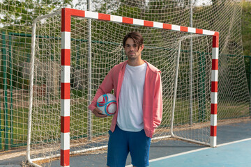 Wall Mural - Man holding a soccer ball under his arm. Close-up image of a person in a pink sweatshirt holding a white, red, and blue ball in front of a soccer goal on a blue court with yellow lines and nets on..