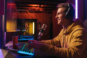 Wall Mural -  Excited caucasian gamer streamer with headphones playing computer pc video at his living room, recording live stream using microphone, drinking energy drink. Cyber sport and social media concept