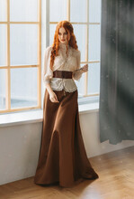 Red-haired Woman In Vintage Dress Stands At Large Classic Window Waiting Love. Clothing Costume Countess Old Style White Blouse, Brown Long Skirt. Curly Red Hair. Redhead Girl Princess 1800s Stylish