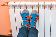Close-up Of Legs Dressed In Warm Socks On A White Central Heating Battery.