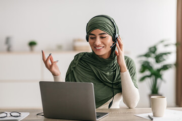 Wall Mural - Positive young Arab woman in hijab and headphones video chatting, having online conference on laptop at home