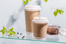 Coffee To Go. Two Disposable Eco-friendly Cardboard Cups, Ground Coffee In A Glass Jar On A Glass Shelf On Blue Background With Green Branches And Copy Space. Takeaway Coffee Concept. Soft Focus Stile