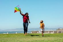 African American Father Holding Kite While Running By Son On Grassy Promenade Against Blue Sky