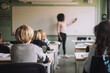 © Maskot - Rear view of students sitting at desk while teacher teaching in classroom