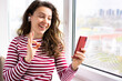 © Parten - Happy young woman using mobile phone for video calling conversation, showing ok okay gesture, wearing red striped t-shirt standing near window indoors. Using 5g wireless internet connection