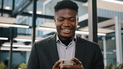 Happy smiling african guy american man businessman boss employee holding modern wireless device texting message on smartphone using social media apps browsing internet playing mobile game at office