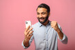 © Vadim Pastuh - Astonishment Indian man using a smartphone isolated on pink background, hispanic guy looks at the screen with mouth opened holding phone, excited with good deal or unexpected news