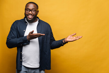 Smiling cheerful young African American man points both hands aside looking at the camera with cheerful wide toothy smile, black guy presenting novelty, advertising, isolated on gray wall