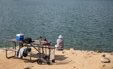 Winchester, CA, USA - April 11, 2022: Skinner Lake. In Front Of Dark Blue-gray Water, Fisher Sits, Back To Us, On Sandy Rocky Shoreline With His Tools Exposed On Old Wooden Bench.