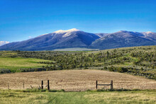 A Farm Gate In The Foreground Frames A Distant Snow-covered Mountain In Montana, Viewed Beyond Hilly Fields Of Brown And Green.