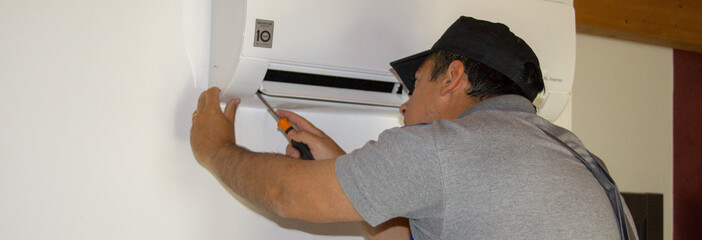 Wall Mural - Plumber on a ladder who, with a screwdriver in hand, carries out maintenance on an air conditioner during the summer. 