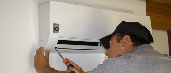 Wall Mural - Plumber on a ladder who, with a screwdriver in hand, carries out maintenance on an air conditioner during the summer. Horizontal banner 