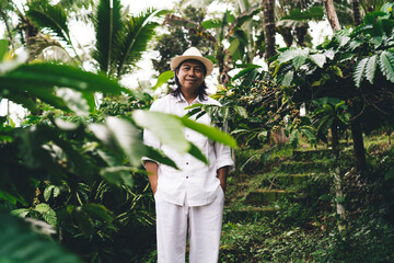 Wall Mural - Portrait of successful Balinese male in hat looking at camera during daytime at seedling field with coffee bush, adult farmer posing while visiting countryside caffeine plantation in Indonesia