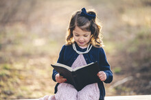 Cheerful Little Girl Sitting On A Park Bench And Reading A Book