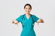 Covid-19, healthcare workers, pandemic concept. Smiling pleasant asian female doctor, therapist or physician in scrubs with stethoscope, pointing fingers down, show clinic banner