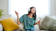 Young Asian Woman Listening To Music On Couch In Living Room At Home. Happy Asia Female Using Mobile Smartphone, Wearing Headset And Sitting On Sofa