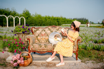 Wall Mural - Cheerful pretty young woman with hair tied by handkerchief reading a book with cute small prairie dog sitting on vintage sofa relaxing in beautiful flower garden. Asian people.