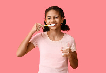 Sticker - African-American teenage girl brushing teeth with glass of water on pink background