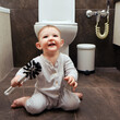 Happy toddler baby boy is playing in the toilet room with a brush. Smiling child plays on the brown floor in the beige bathroom