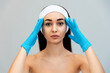 © _KUBE_ - Portrait of a young caucasian woman with a bandage on her head, leaning her gloved hands against her foreground. Gray background. The concept of professional cosmetology and plastic surgery