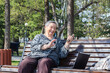 © Kris Zarzh - Attractive senior woman sitting on bench using smartphone and computer for remote online chat.