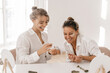 © Look! - Happy young caucasian ladies in good mood use face cream sitting in studio. Blondes wear white shirts laugh together. Day with skin care and health.