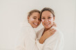 Positive two young caucasian women smile teeth standing inembrace on white background. Blondes wear bathrobes and patches during spa treatments. Concept of natural cosmetics, wrinkle smoothing