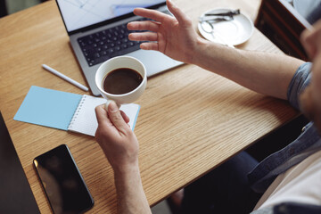 Wall Mural - Close up of white cup of black coffee in male hand. Caucasian man in cafe with laptop. Top view.