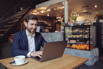 Wall Mural - Caucasian attractive man sitting at table and texting on laptop in luxury cafe. Freelancing work.