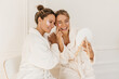 © Look! - Pretty caucasian young women wear bathrobes admire themselves looking in mirror on white background. Blondes use eye patches for dark circles. Beauty and personal care, skin and face care concept