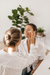 © Look! - European girl applies skin care cosmetics on face of happy adult woman sitting in light room. Blondes wear white shirts. Beauty treatment, morning ritual
